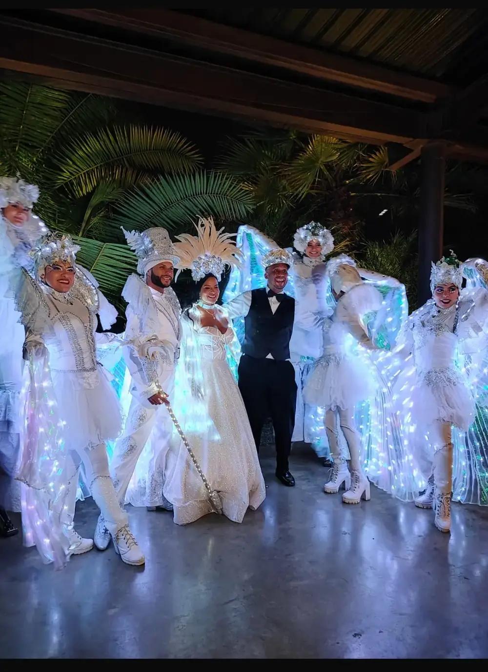 Pareja de novios rodeada de zanqueros con trajes blancos y alas luminosas en una noche de boda
